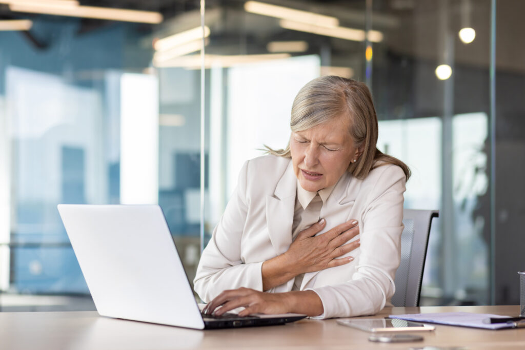 Heart attack chest pain, senior mature businesswoman in business suit working inside office at workplace with laptop, having severe heart pain holding hand to chest